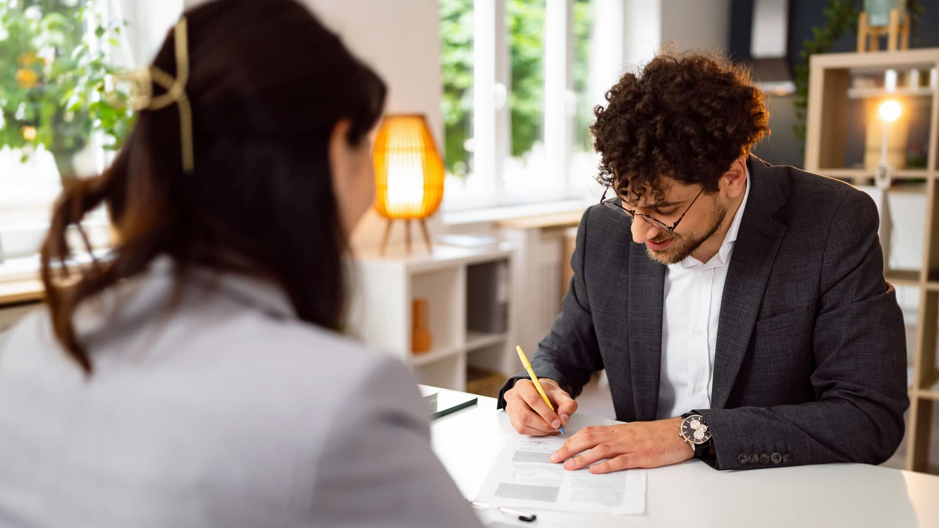 Blick über die Schulter einer Person auf einen jungen Mann, der am gleichen Tisch sitzt und Dokumente unterschreibt