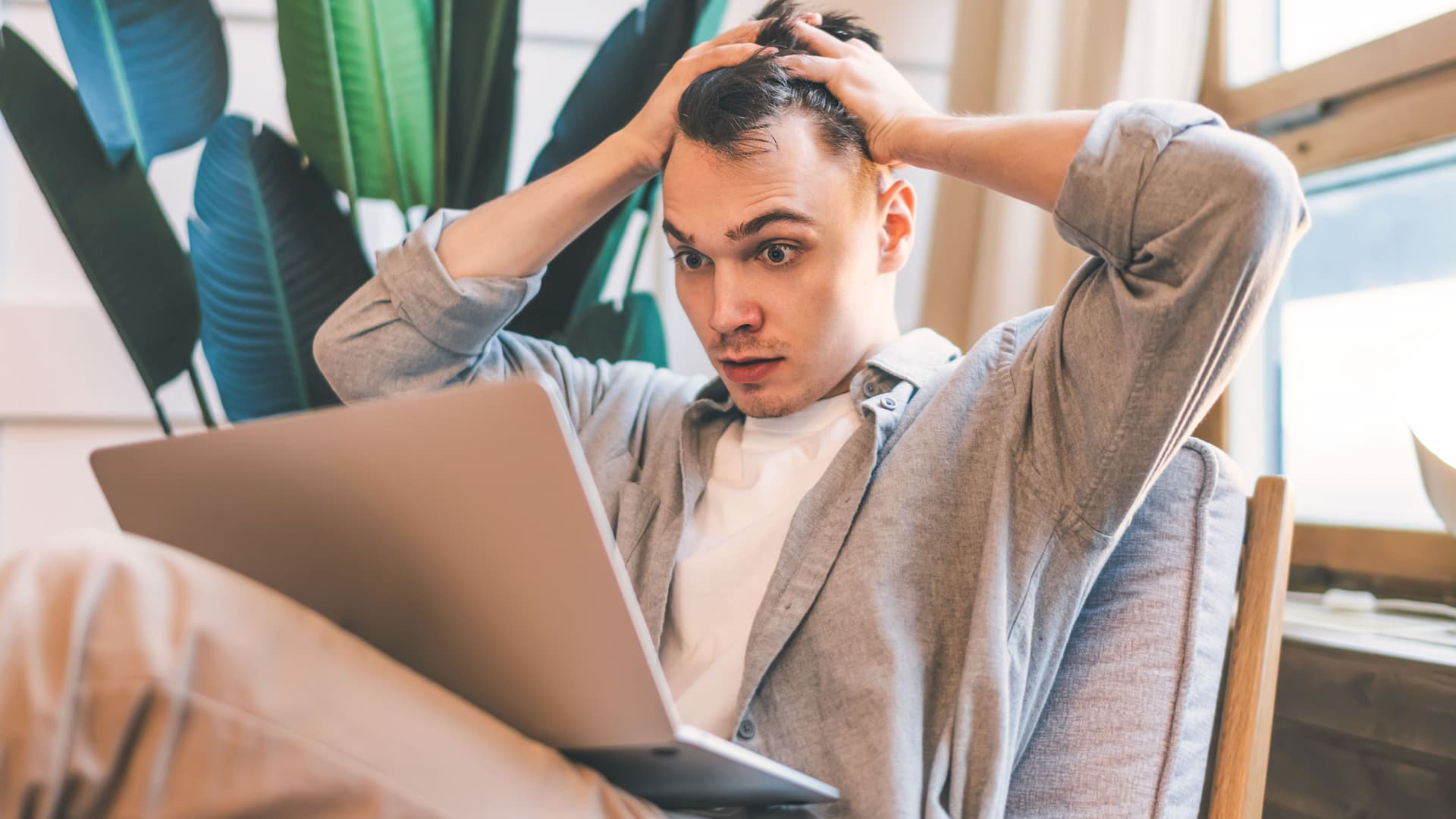 Überraschter junger Mann schaut auf Laptopbildschirm und rauft sich die Haare. Er sitzt in einer modernen Wohnung in einem Sessel.