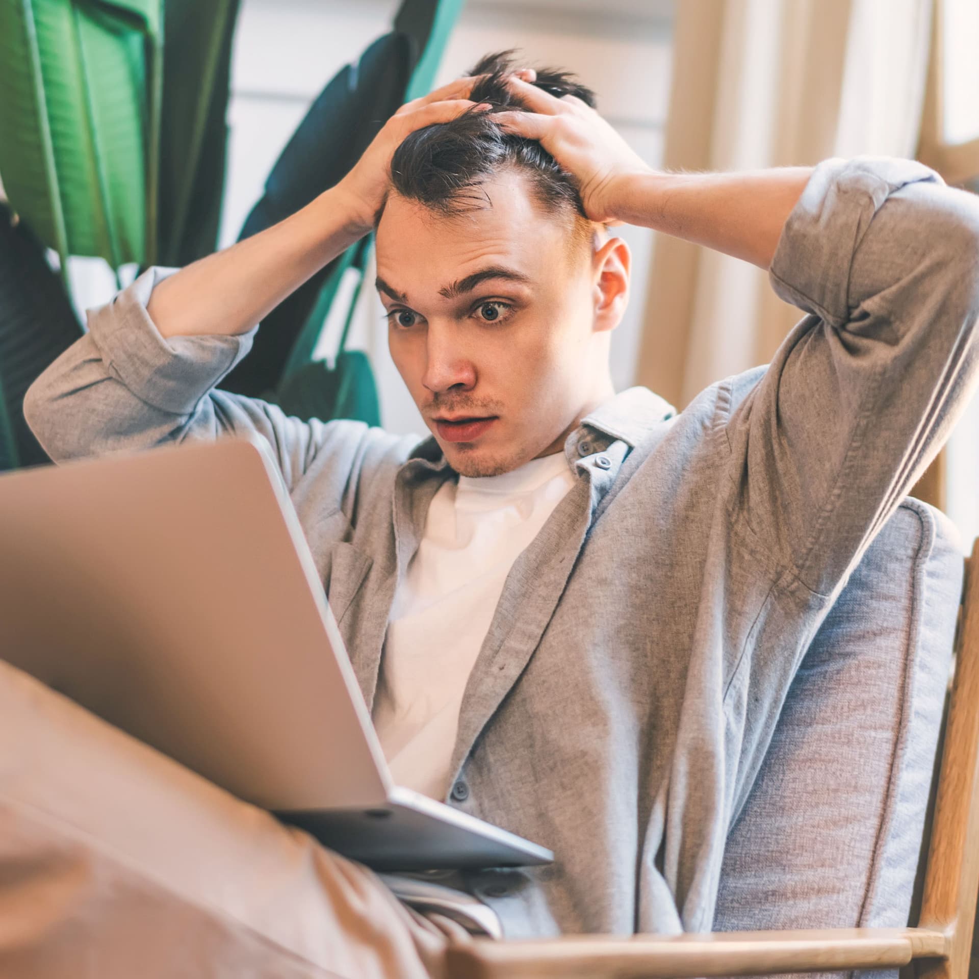 Überraschter junger Mann schaut auf Laptopbildschirm und rauft sich die Haare. Er sitzt in einer modernen Wohnung in einem Sessel.