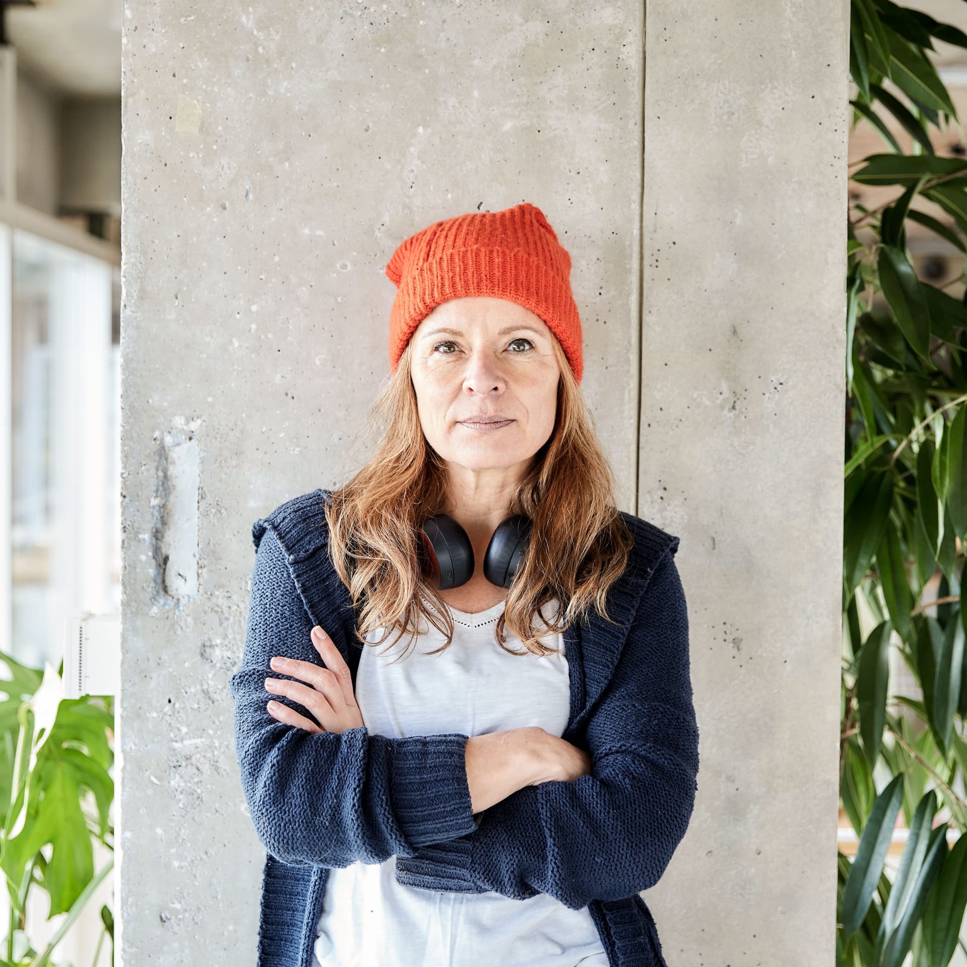 Ältere Frau mit verschränkten Armen steht an einer Säule. Um den Hals trägt sie Kopfhörer.