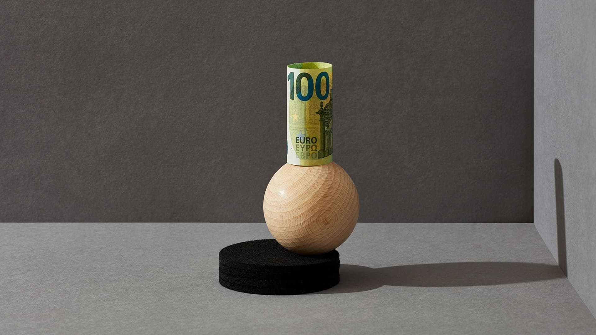 Eine gerollte 100-Euro-Banknote steht senkrecht aufgerichtet auf einer Holzkugel vor grauem Hintergrund.