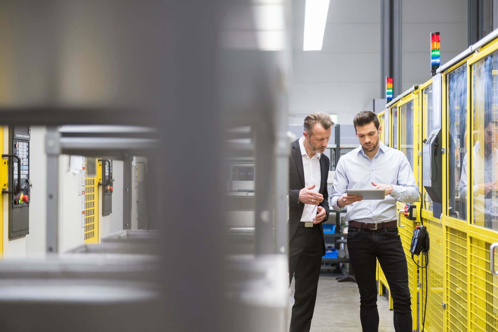 Zwei Männer in Anzug stehen in einer Fabrik mit Industrierobotern und gucken zusammen auf ein Tablet.