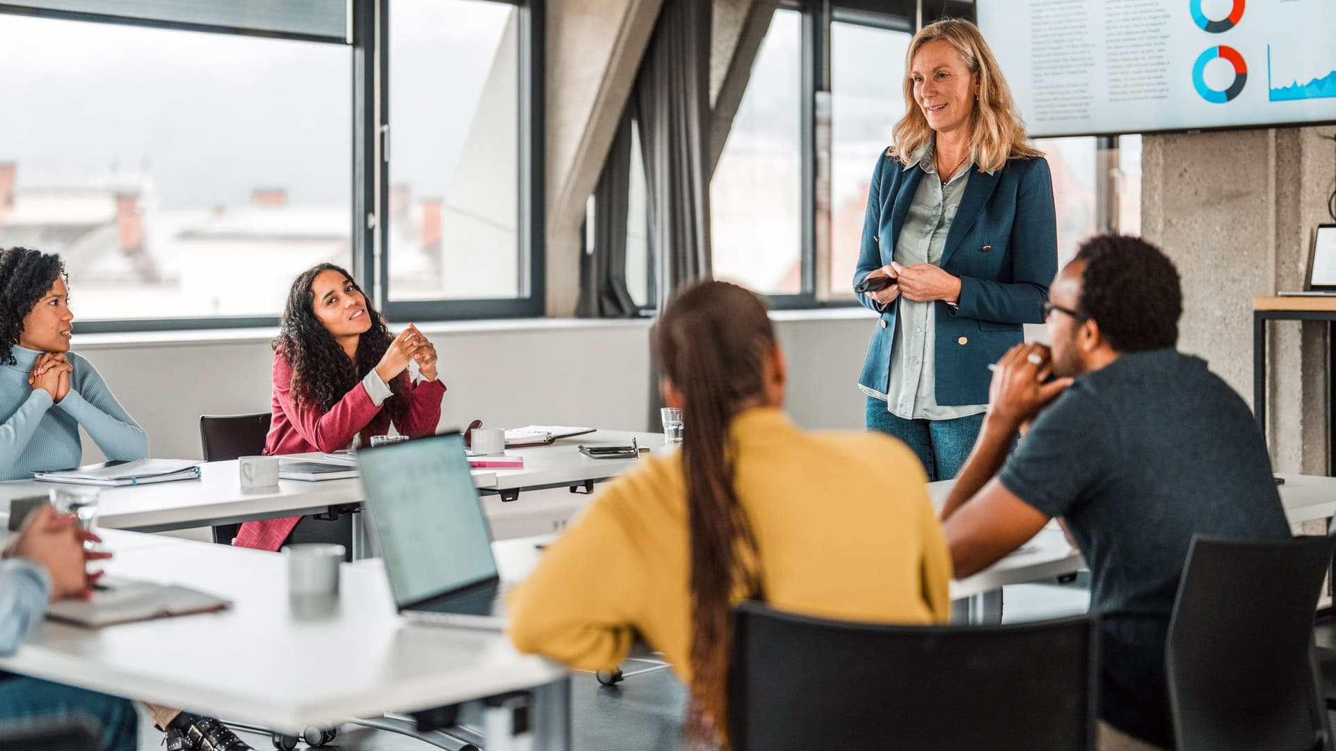 Frau präsentiert Geschäftszahlen vor einer Gruppe diverser Kolleg:innen in einem modernen Konferenzraum. Menschen sitzen am Tisch, hören aufmerksam zu und diskutieren.