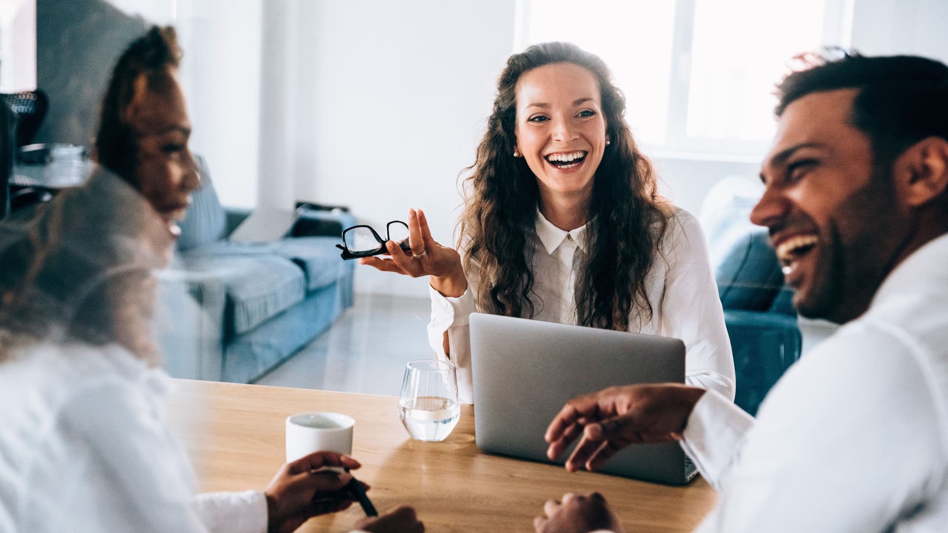 Zwei Frauen und ein Mann sitzend lachend an einem Bürotisch. Eine Frau hat einen aufgeklappten Laptop vor sich auf dem Tisch.
