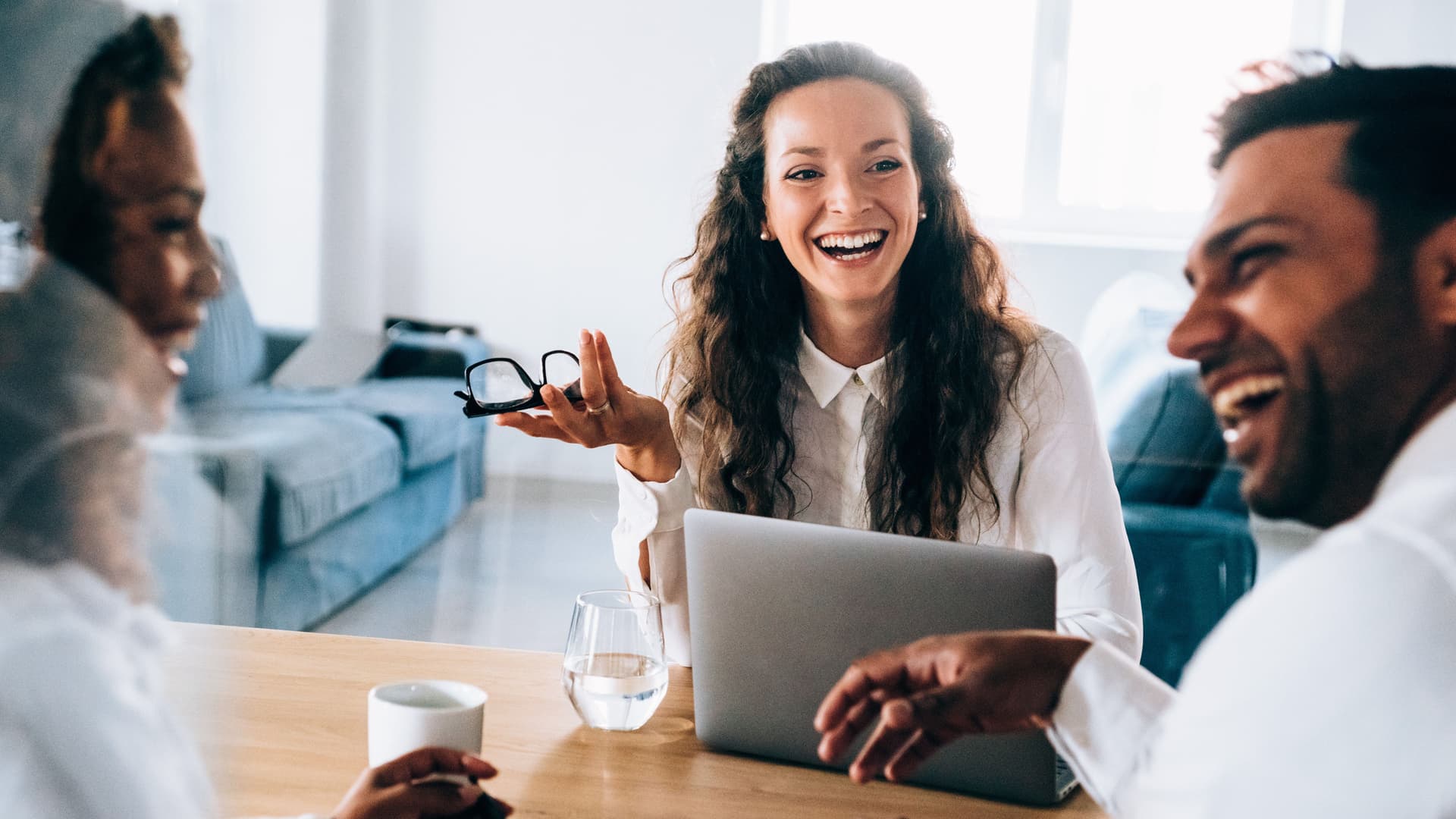 Zwei Frauen und ein Mann sitzend lachend an einem Bürotisch. Eine Frau hat einen aufgeklappten Laptop vor sich auf dem Tisch.