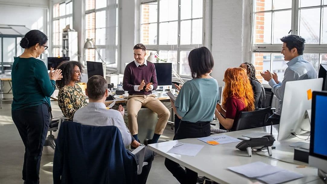 Bürokolleg:innen sitzen und stehen im Kreis zwischen Arbeitsplätzen und klatschen für eine Kollegin