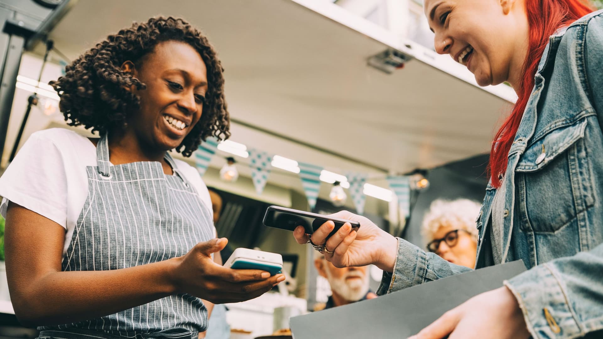Eine junge Frau zahlt kontaktlos mit Handy. Die Kellnerin steht vor einem Foodtruck und hält ihr ein Kartenlesegerät entgegen.