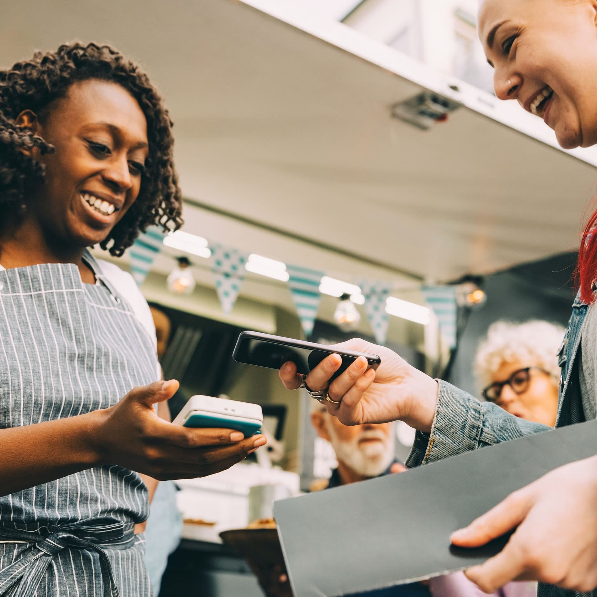 Eine junge Frau zahlt kontaktlos mit Handy. Die Kellnerin steht vor einem Foodtruck und hält ihr ein Kartenlesegerät entgegen.