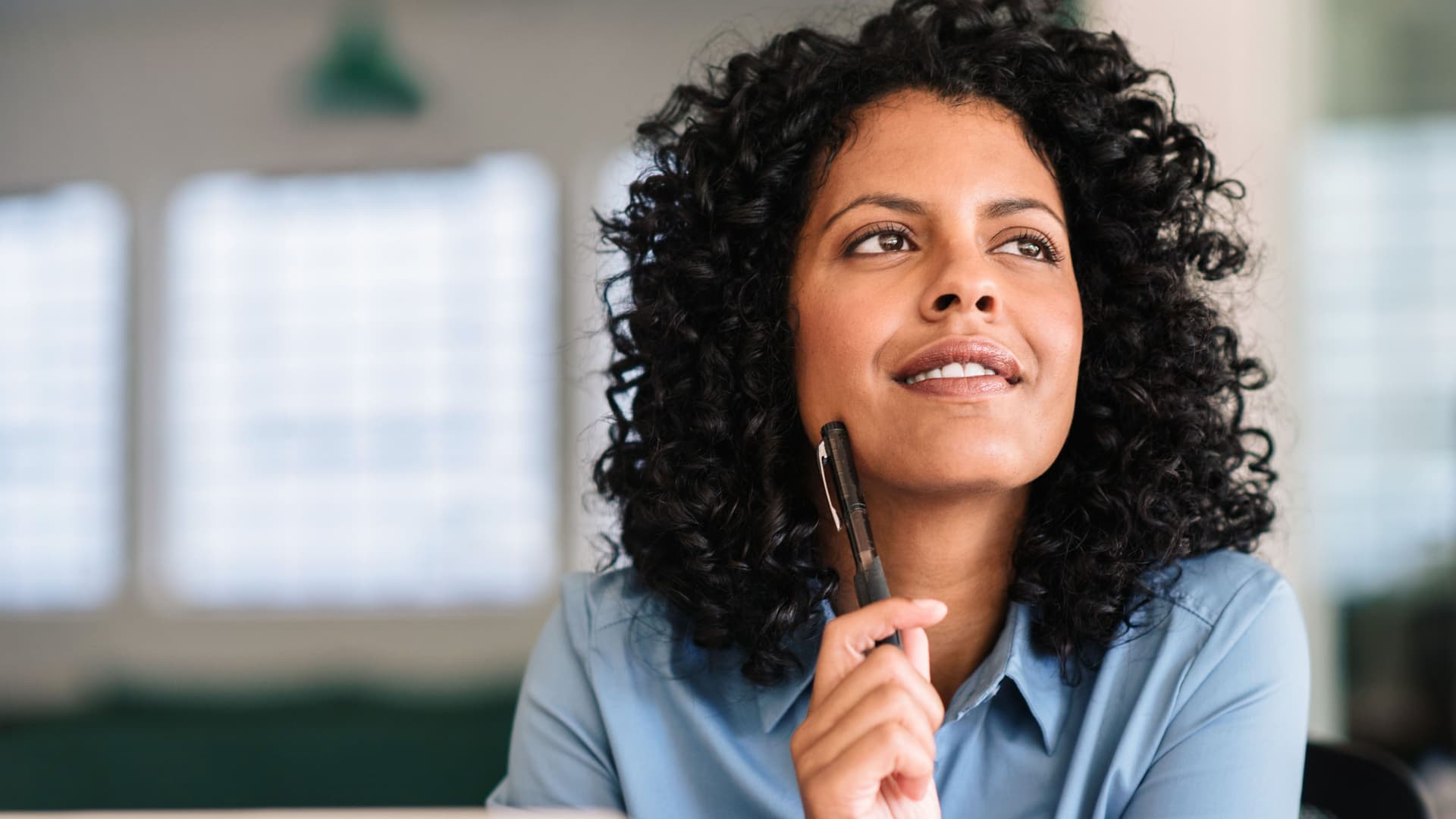 Selbstständig machen Lächelnde junge Frau guckt tief in Gedanken nach oben und hält einen Stift in der Hand. Sie sitzt an einem Schreibtisch in einem Büro mit vielen Fenstern.