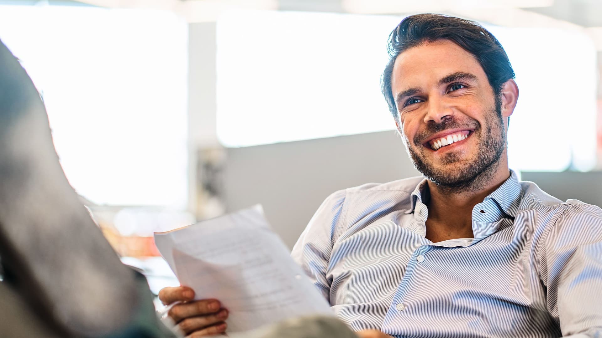 Lächelnder junger Mann, der Papiere liest, während er mit den Füßen hoch auf seinem Schreibtisch in einem großen, modernen Büro sitzt.