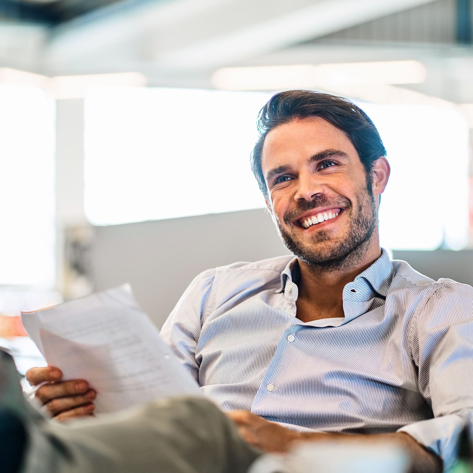 Lächelnder junger Mann, der Papiere liest, während er mit den Füßen hoch auf seinem Schreibtisch in einem großen, modernen Büro sitzt.