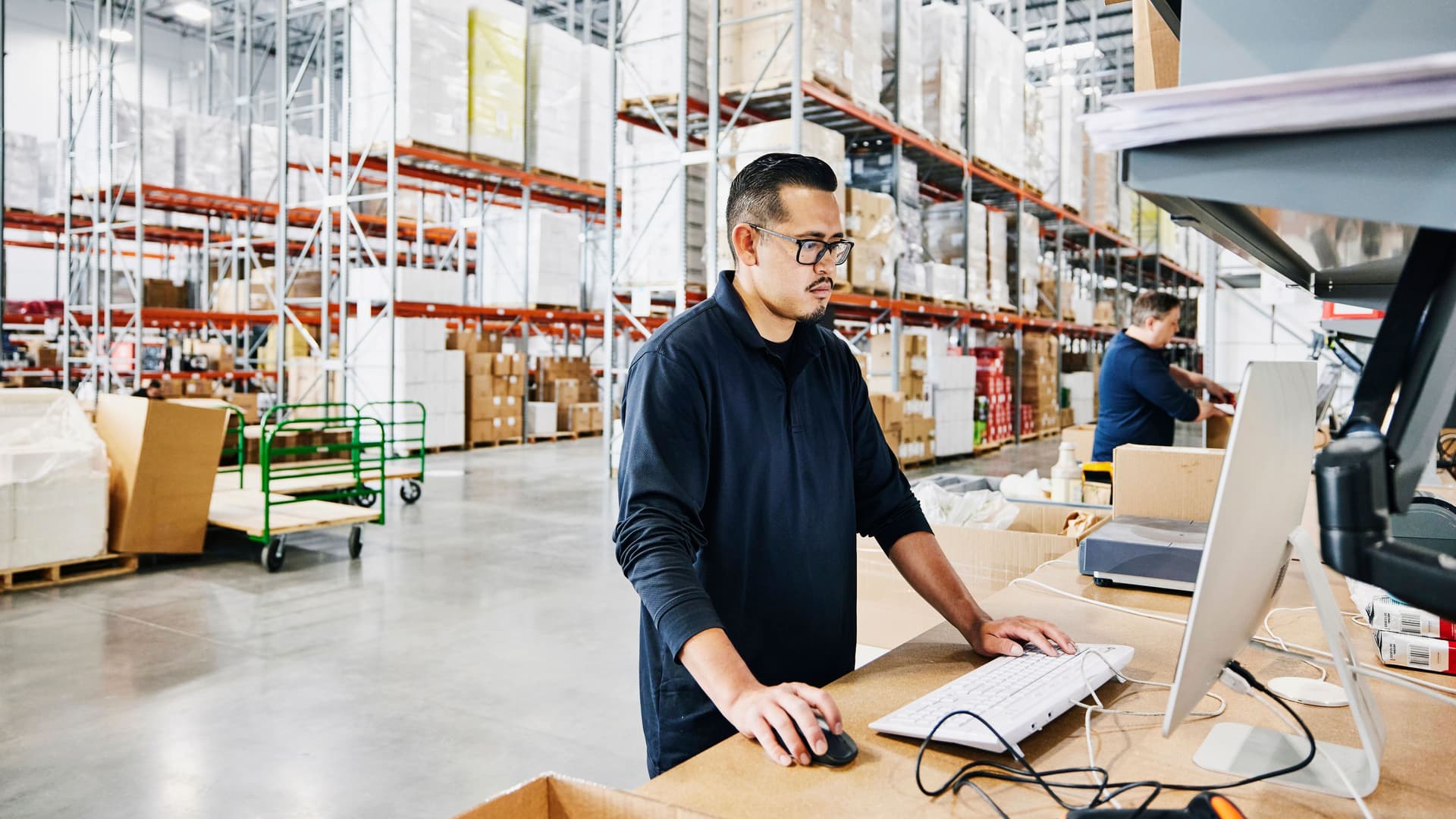 Lagerarbeiter überprüft Bestellungen an einem Computerarbeitsplatz in einem Lager. Im Hintergrund packt ein weiterer Arbeiter Kisten.