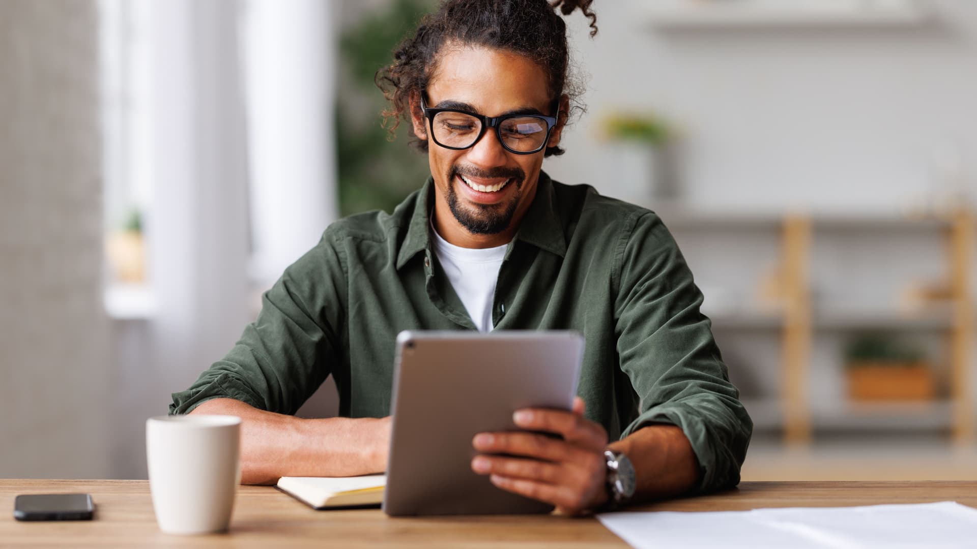 Junger Mann sitzt an einem Holztisch. Er schaut auf sein Tablet und lächelt.