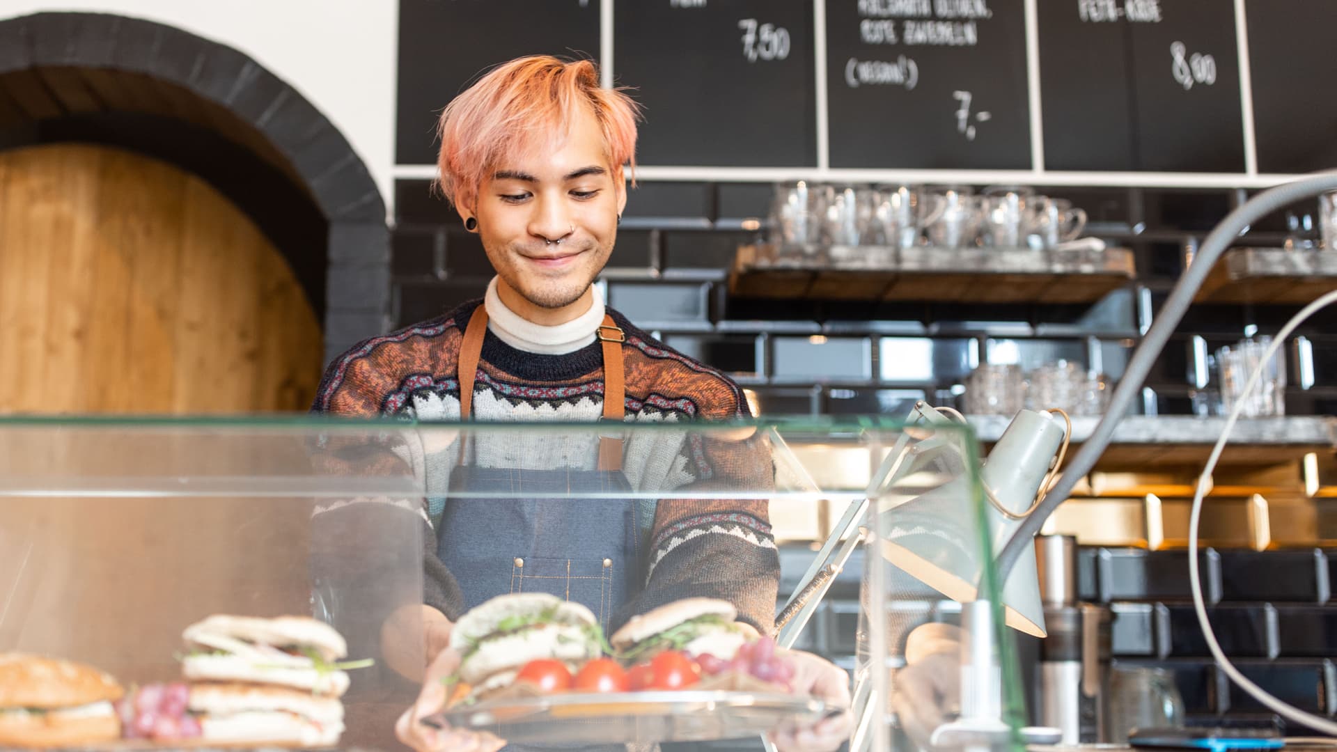 Ein Mann mit rosa gefärbten Haaren arbeitet in einem Cafe und befüllt die Theke mit Paninis.