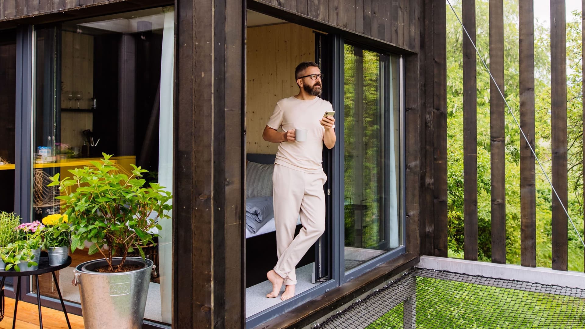 Ein Mann steht barfuß mit einer Tasse Kaffee und einem Smartphone am Fenster eines Tiny Houses und blickt dabei in die Natur.