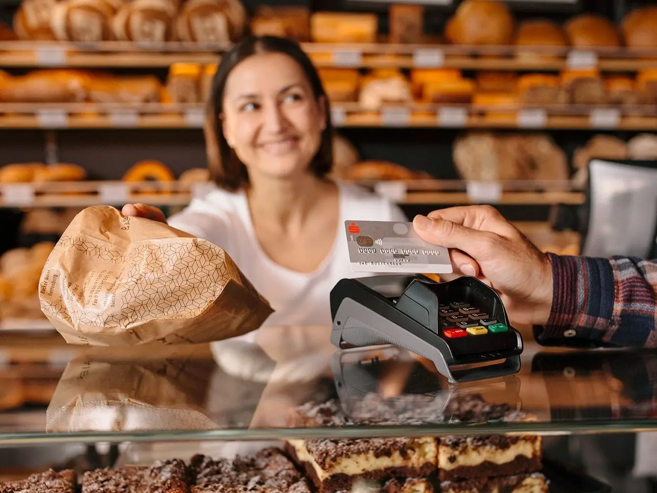 Eine männliche Hand hält in einer Bäckerei seine Sparkassenbankkarte an ein Kartenlesegerät. Im Hintergrund sieht man eine Bäckereifachangestellte die eine Brötchentüte neben ihm ablegt.