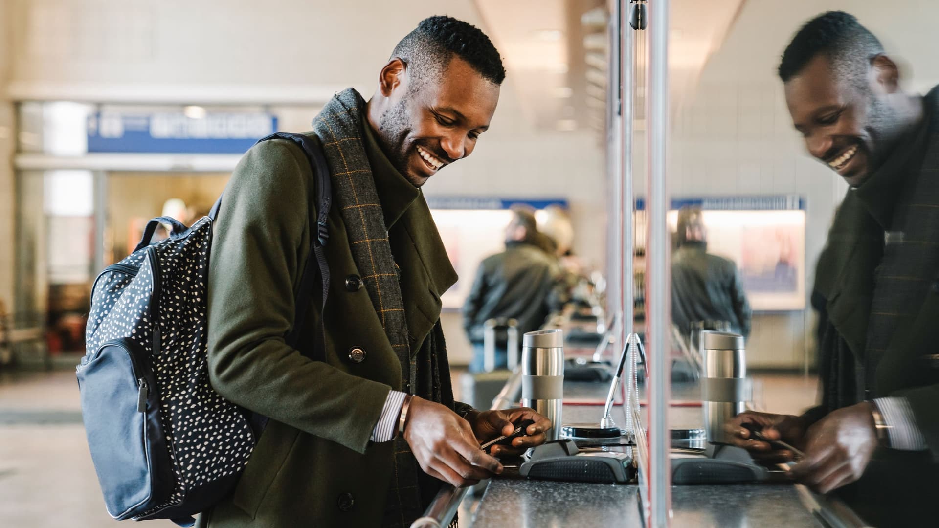 Ein junger Mann zahlt mit einem strahlenden Lächeln sein Ticket am Bahnschalter. Er spiegelt sich in der Glasscheibe.