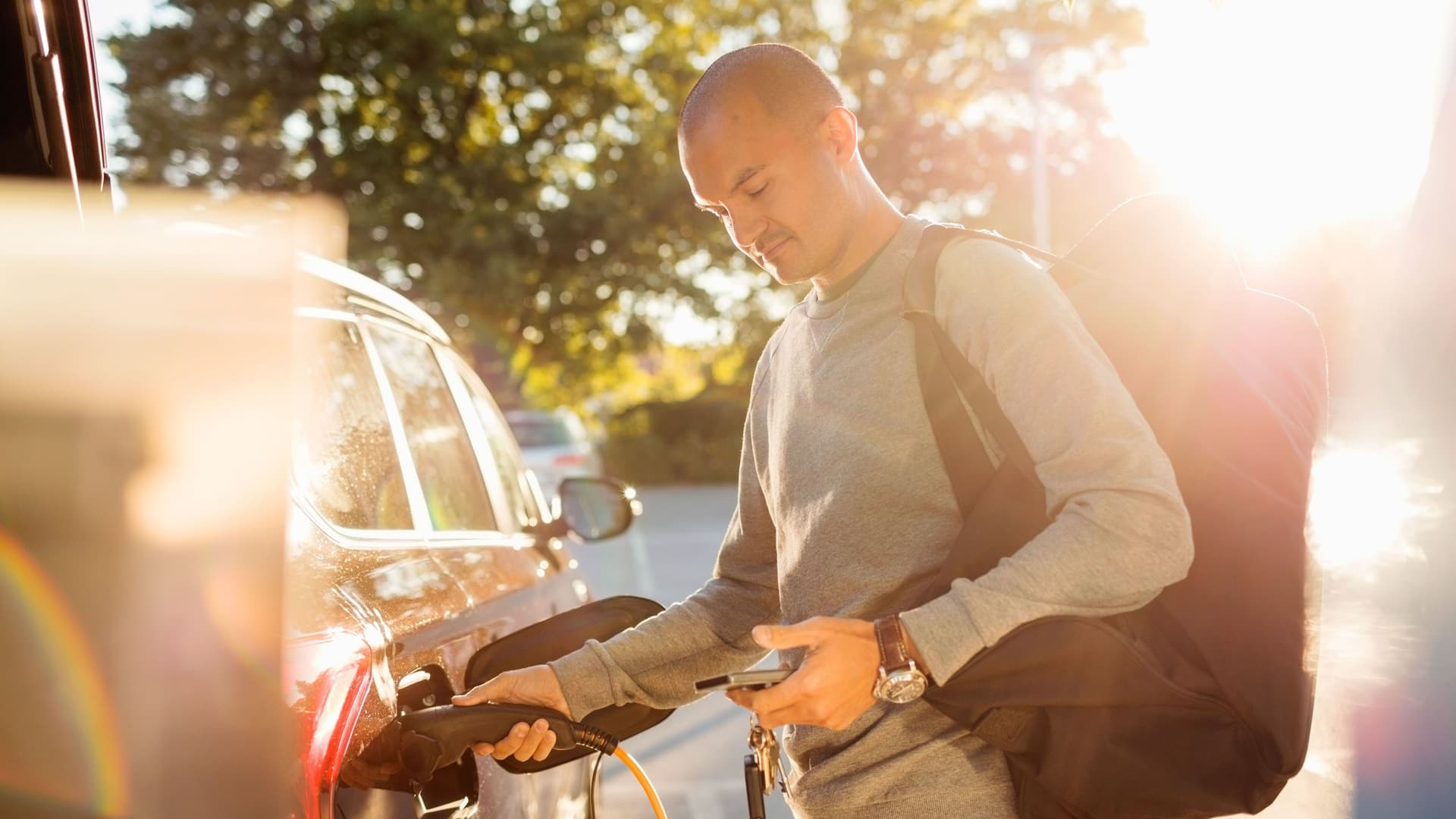 Ein Mann lädt bei Sonnenschein ein Elektroauto auf . Dabei schaut er in sein Smartphone.