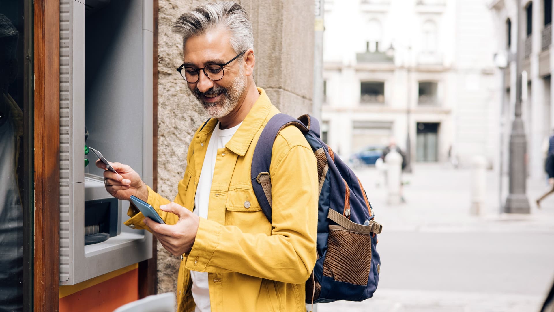 Mann mit grauen Haaren, Brille und gelber Jacke steht lächelnd vor einem Geldautomat. Er schaut auf sein Handy.