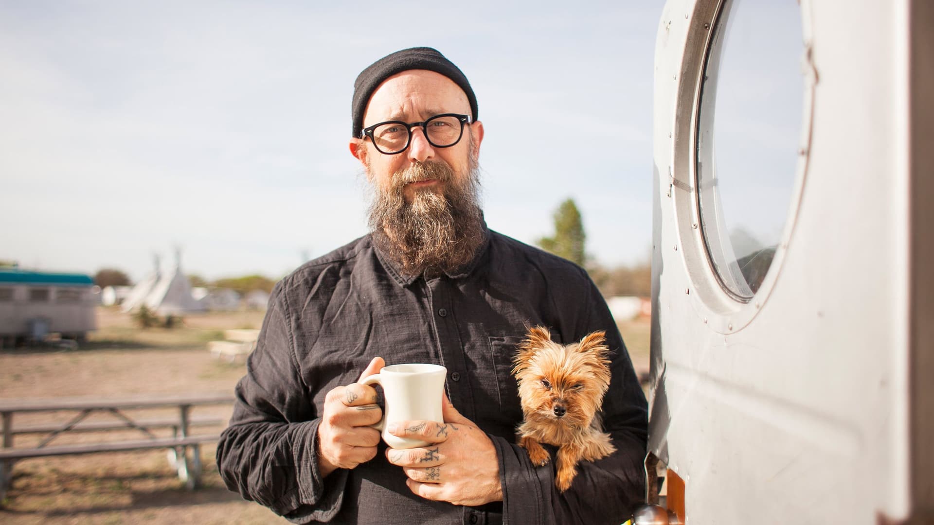 Ein bärtiger Mann mit schwarzer Mütze und Brille steht vor einem Wohnwagen, hält eine weiße Tasse in der Hand und trägt einen kleinen Hund auf dem Arm. Im Hintergrund sind ein Campingplatz und Tipi-Zelte zu sehen.