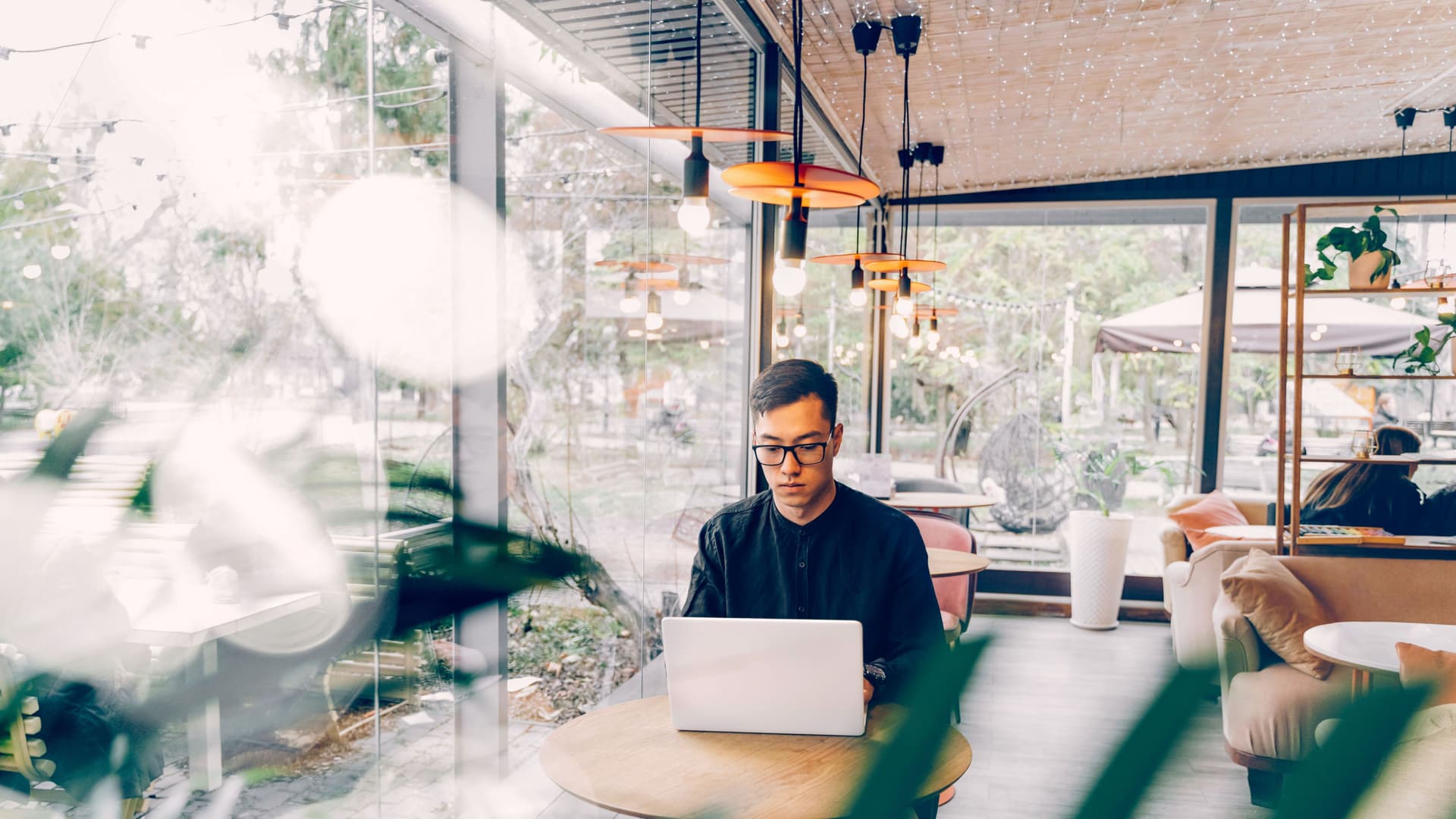 Mann mit Laptop sitzt an einem kleinen Tisch in einem Cafe vor einer großen Glasfront.