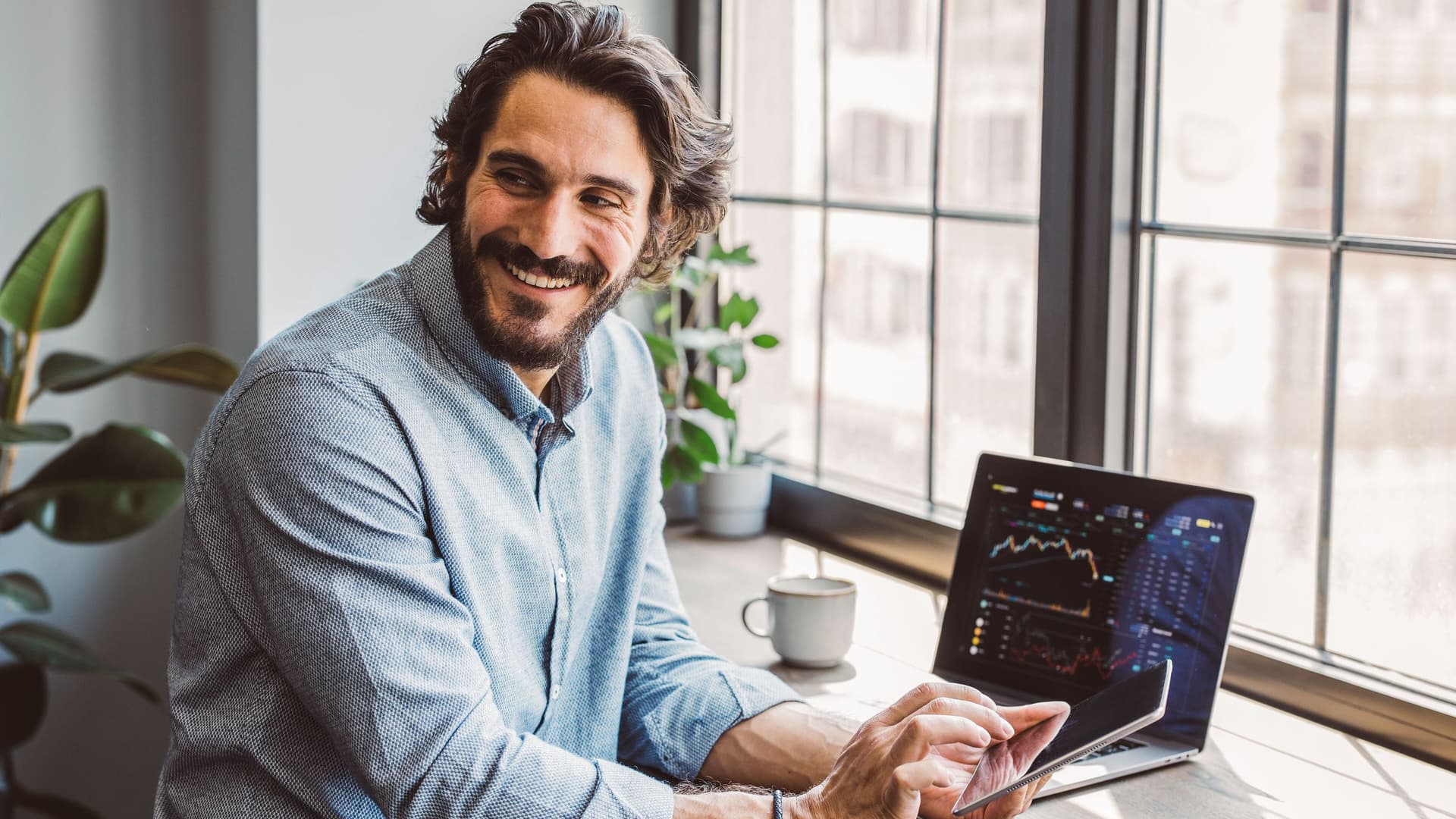 Mann mittleren Alters mit Laptop und Tablet sitzt an einer Fensterfront und lächelt in den Raum.