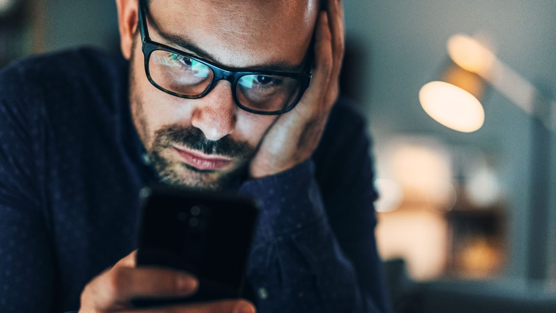 Porträt eines Mannes mittleren Alters. Er hat seinen Kopf aufgestützt und schaut missmutig auf sein Smartphone.