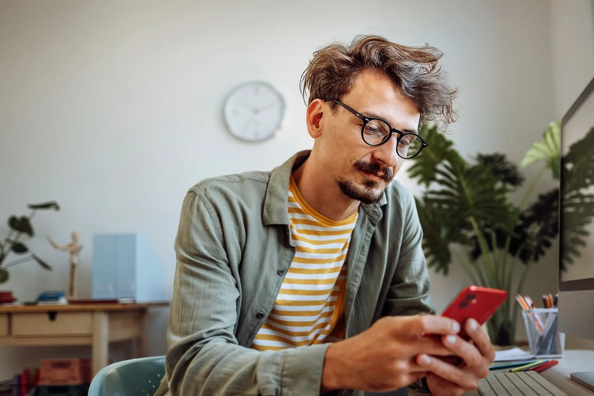 Mann sitzt mit seinem Smartphone vor seinem Computer. Er schaut auf sein Smartphone.