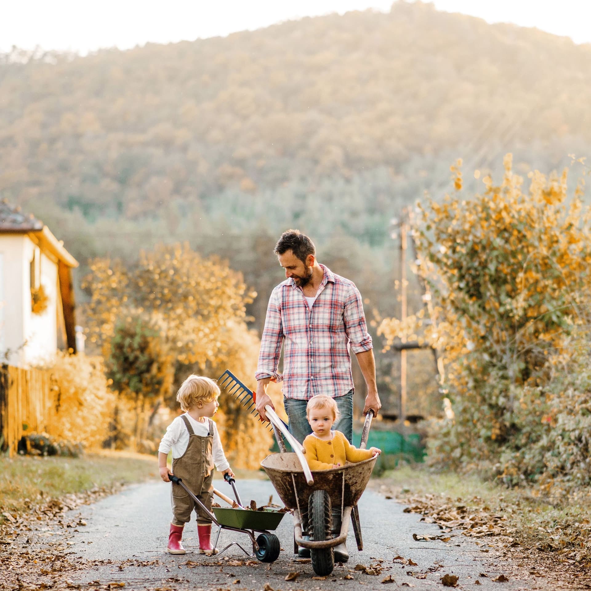 Ein Vater läuft mit zwei Kleinkindern und Schubkarren auf einem ländlichen Weg.