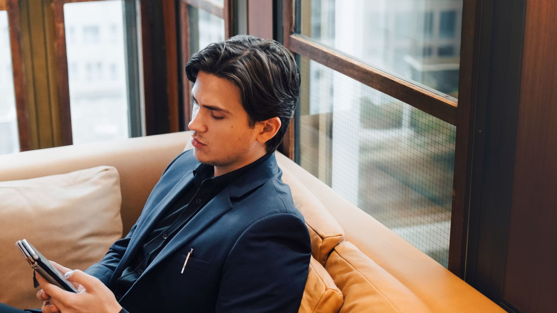 Junger Mann in Anzug sitzt auf einem Sofa vor einer Fensterfront. Er guckt auf das Smartphone in seiner Hand.