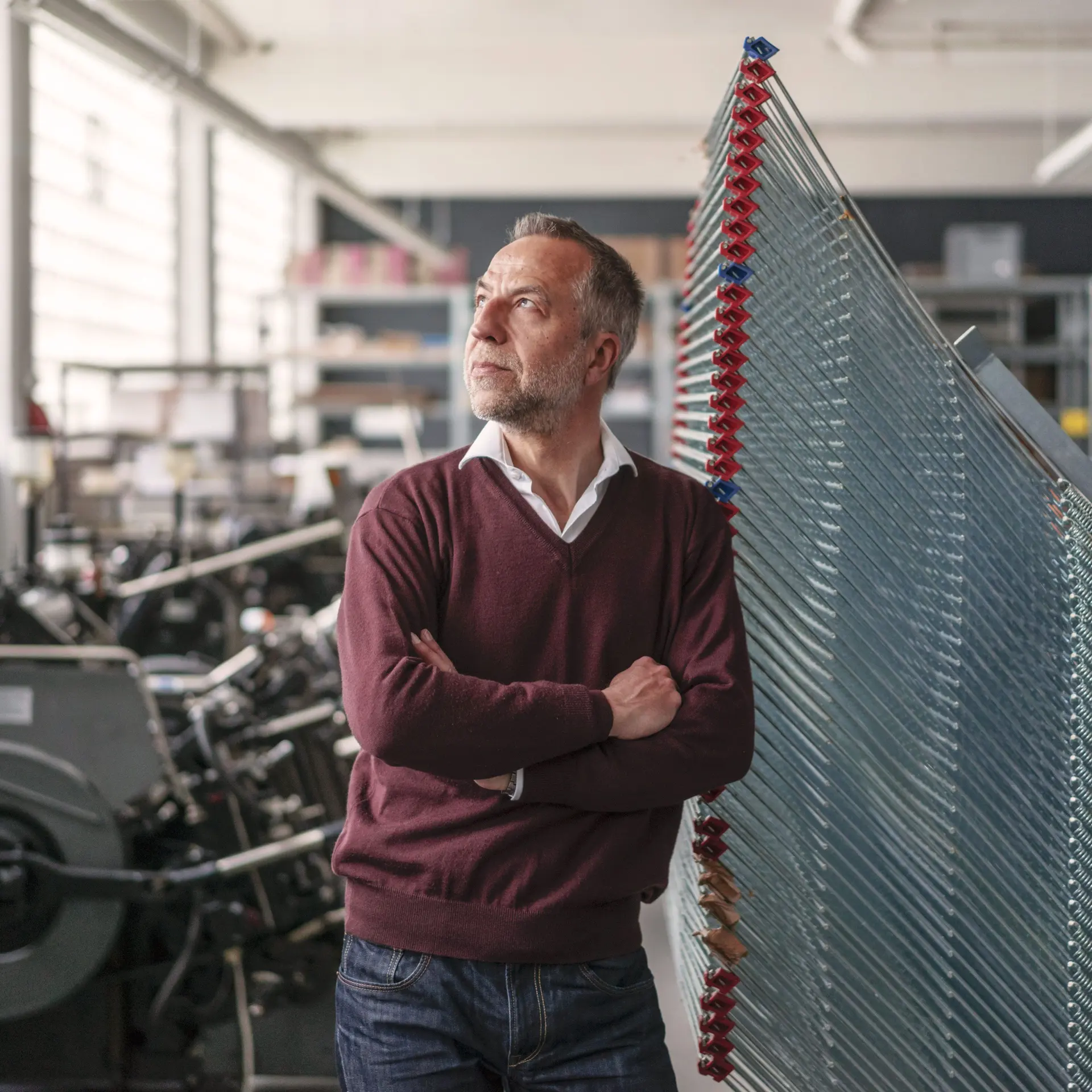 Älterer Mann in einer Druckerei. Er lehnt mit verschränkten Armen an einer Maschine und schaut aus dem Fenster.