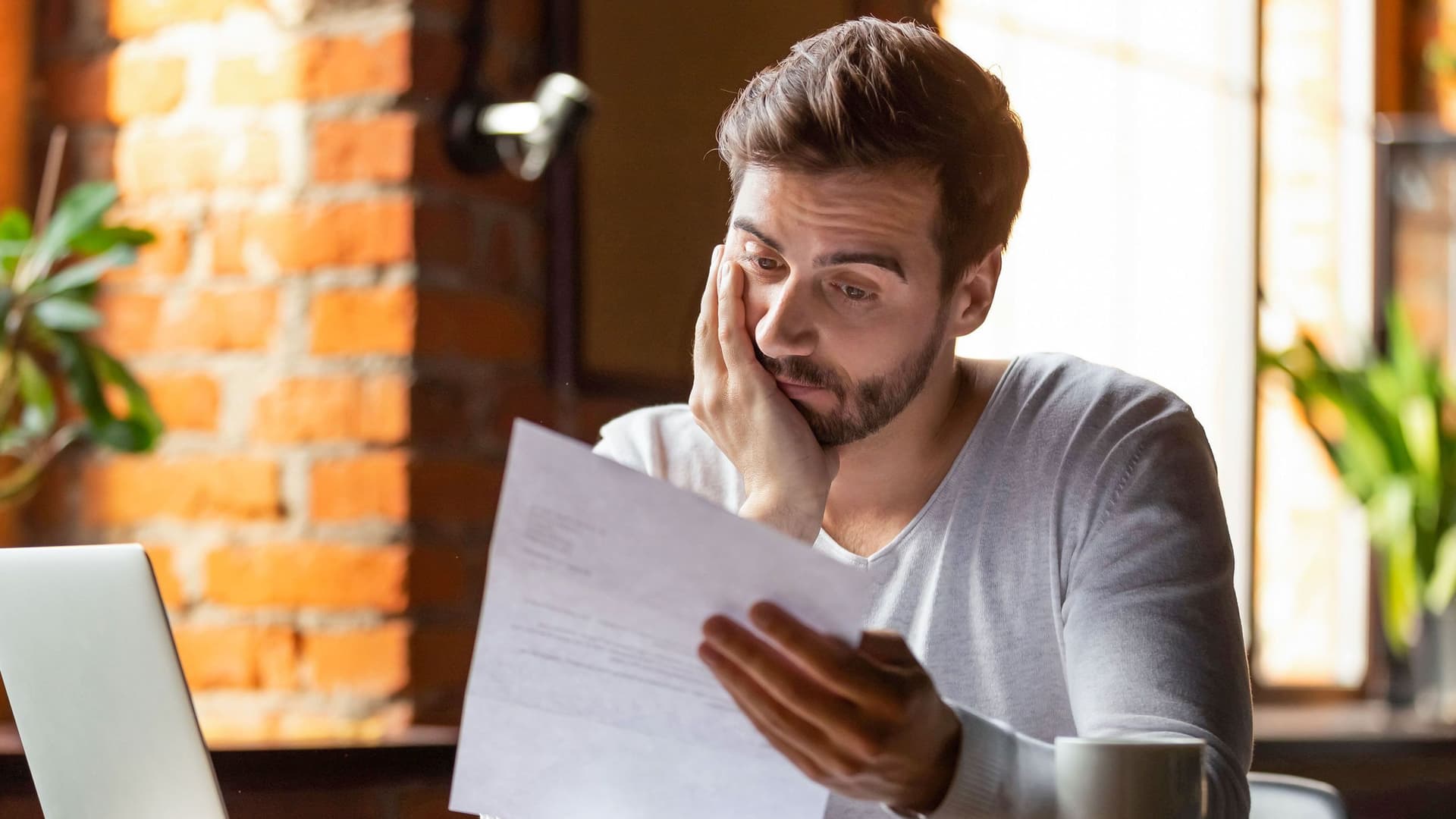 Ein junger Mann sitzt nachdenklich an einem Tisch, vor ihm ein Laptop, eine Tasse und Unterlagen. In einem lichtdurchfluteten Raum mit Backsteinwänden wirkt er besorgt, während er Dokumente betrachtet.
