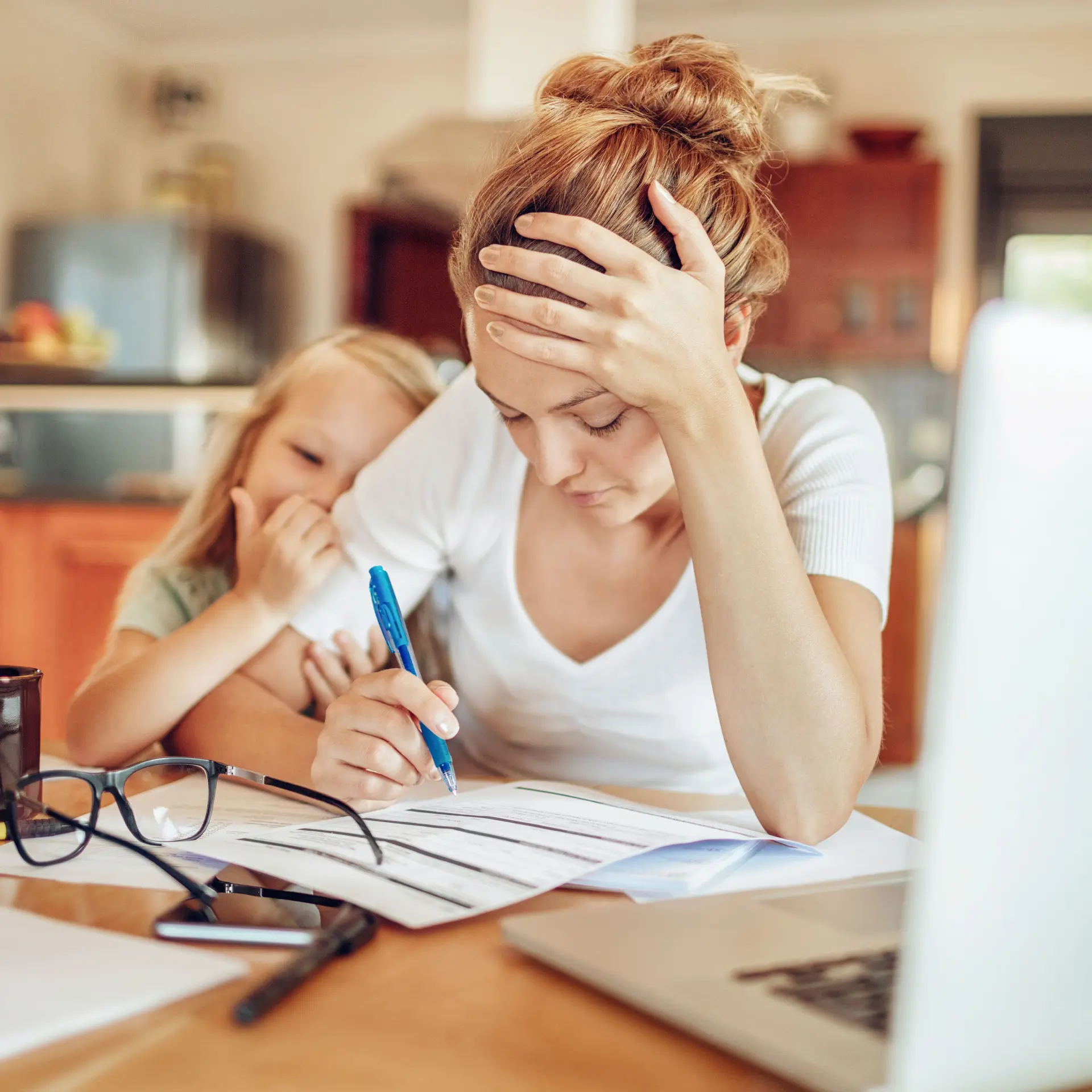 Verzweifelt wirkende junge Mutter mit Kind am Arm sitzt zu Hause vor ihrem Laptop und studiert Unterlagen.