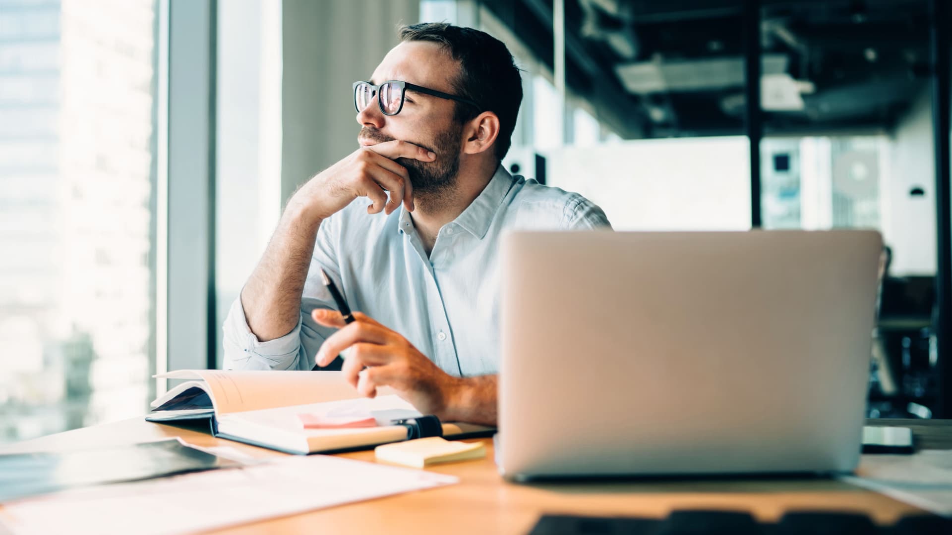 Mann sitzt vor einem Laptop und einem aufgeschlagenem Buch. Er schaut nachdenklich aus dem Fenster eines Büros.