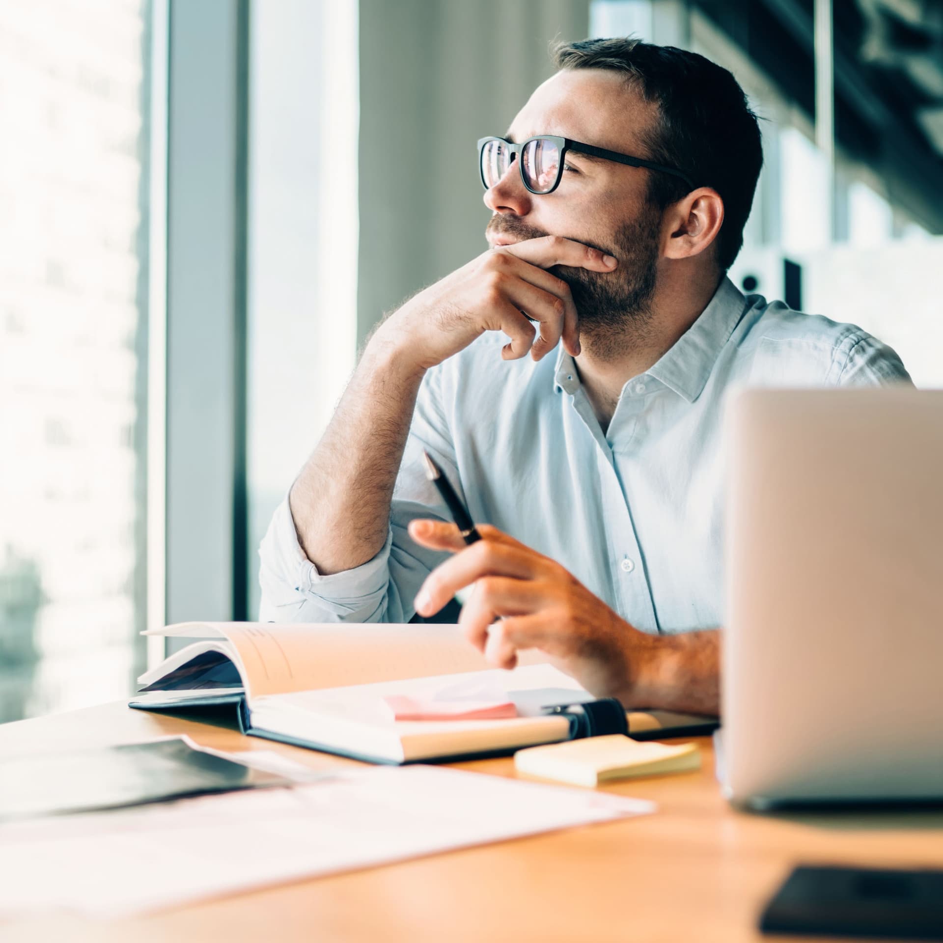 Mann sitzt vor einem Laptop und einem aufgeschlagenem Buch. Er schaut nachdenklich aus dem Fenster eines Büros.