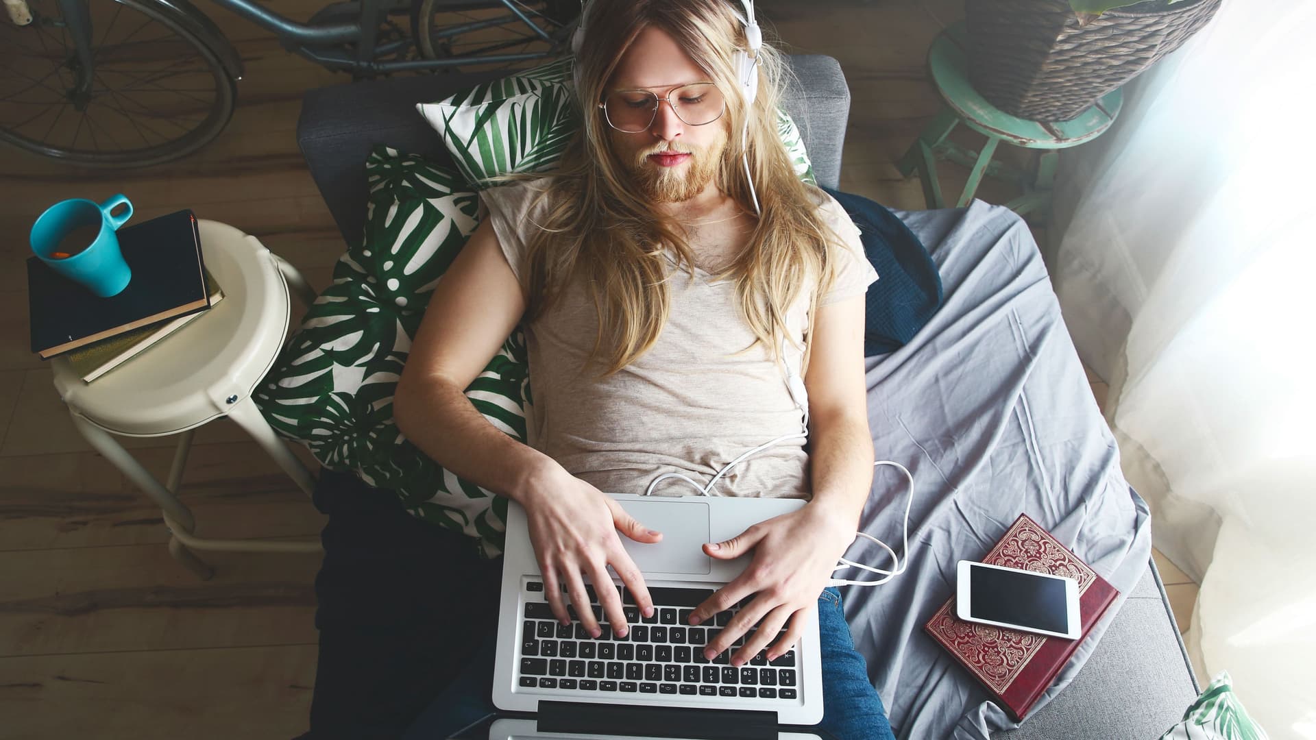 Ein Mann mit langen Haaren und Bart liegt auf einem Sofa. Er trägt Kopfhörer und hat einen Laptop auf seinem Bauch.