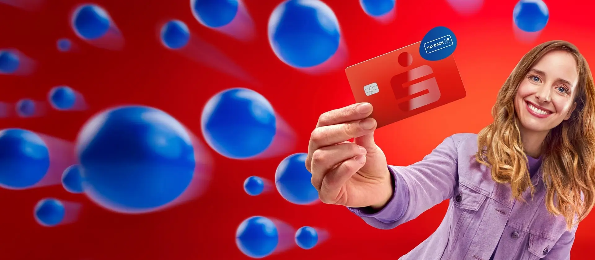 Lächelnde Frau hält eine rote Sparkassen-Debitkarte mit PAYBACK-Logo in die Kamera, vor rotem Hintergrund mit blauen Kugeln