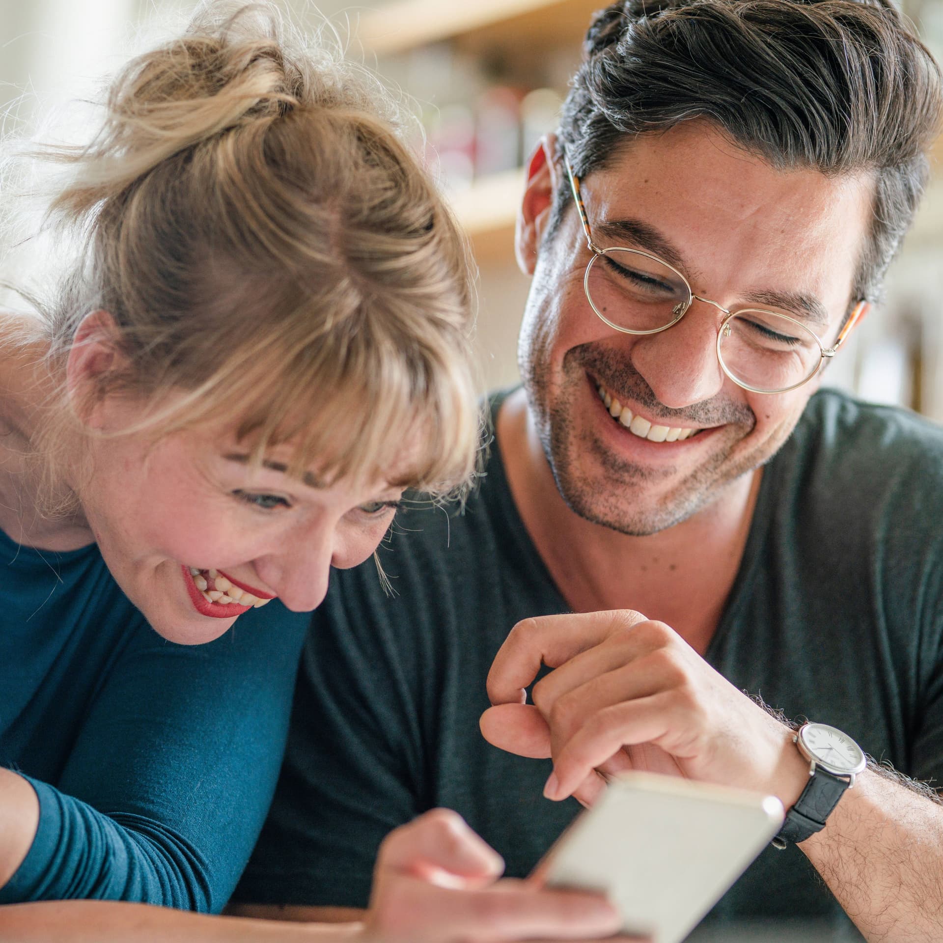 Ein Paar mittleren Alters sitzt zusammen und schaut auf ein Smartphone. Sie lachen ausgelassen.