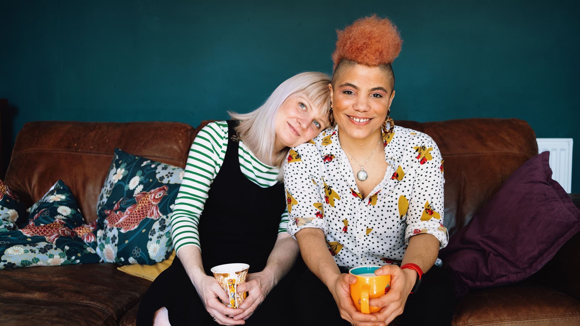 Porträt von zwei Frauen. die nebeneinander mit jeweils einer Tasse in den Händen auf einem braunen Ledersofa sitzen. Die eine Frau hat den Kopf auf die Schulter der anderen gelegt.