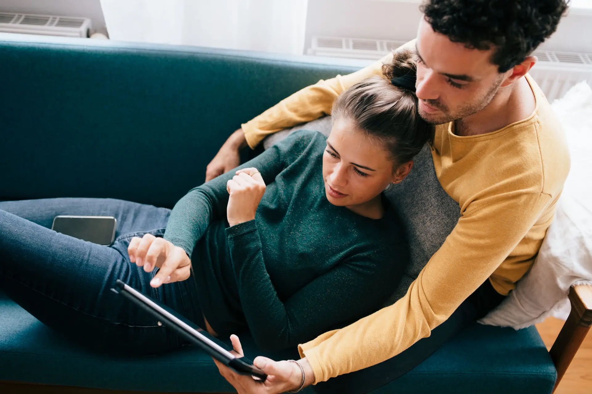 Ein Mann und eine Frau benutzen ein Tablet, während sie sich zu Hause auf dem Sofa liegen. Der Mann umarmt die Frau mit seinem Armen von hinten.