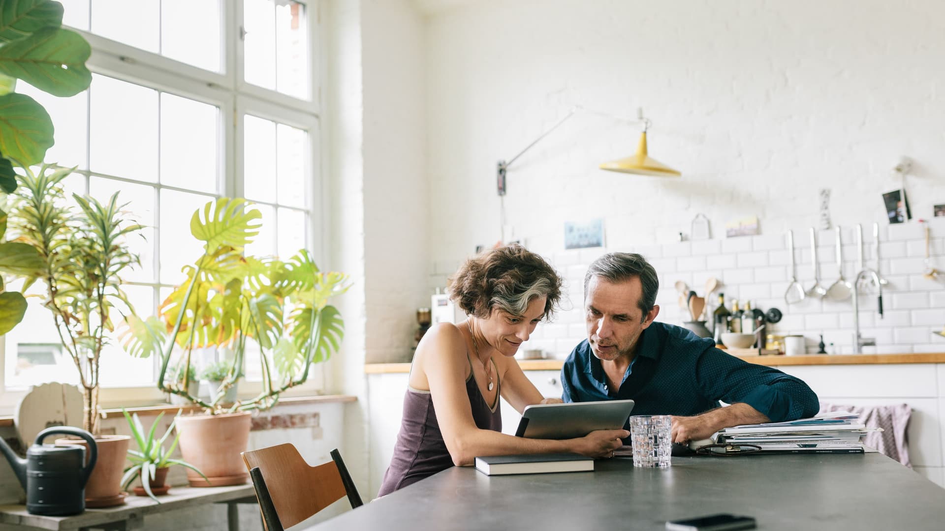 Ein Paar mittleren Alters sitzt eng beieinander in einer großen und lichtdurchfluteten Küche und schaut dabei konzentriert auf ein Tablet.