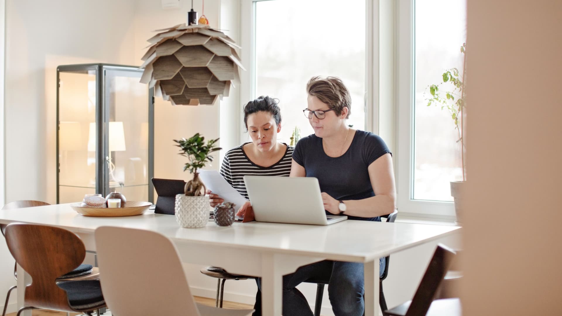 Ein gleichgeschlechtliches Paar sitzt am Wohnzimmertisch und schauen auf Dokumente. Vor den beiden Frauen steht ein Laptop.