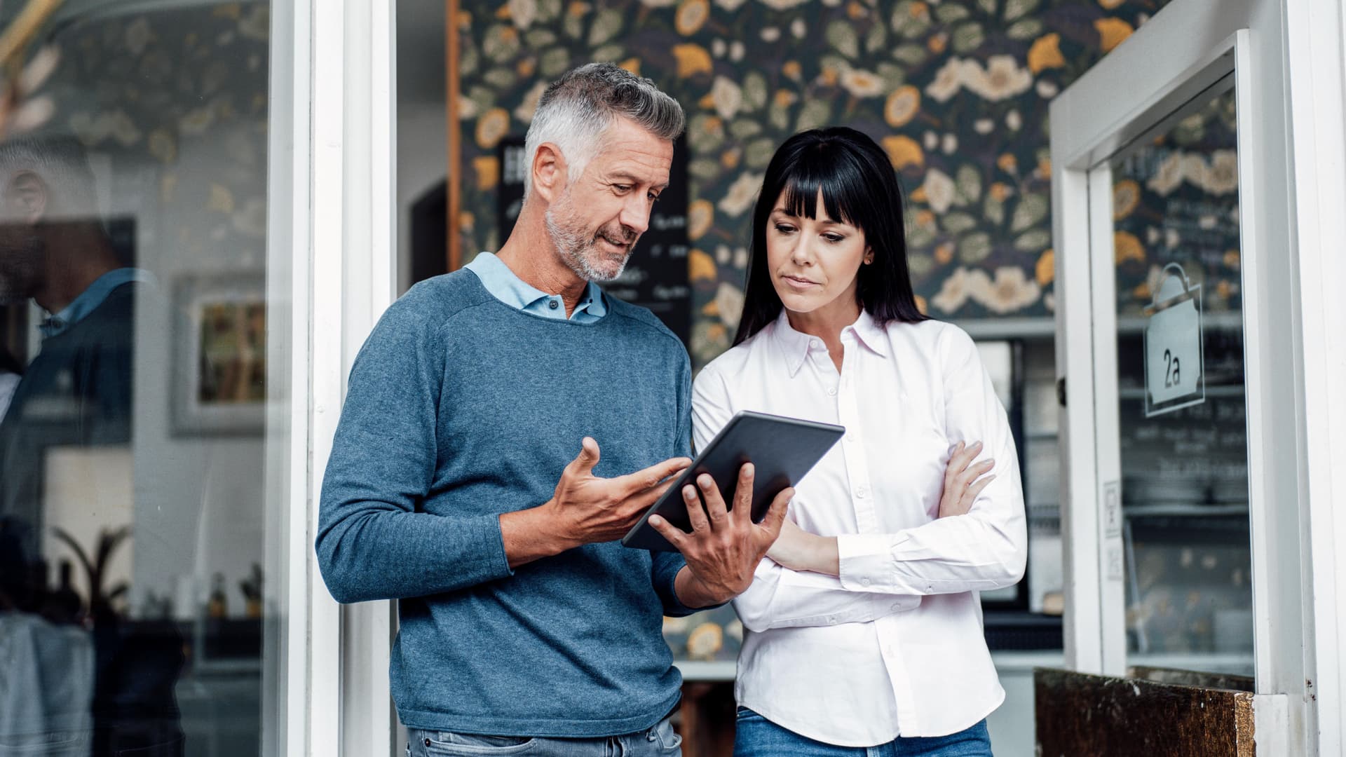 Ein gestikulierender Mann zeigt einer Frau Daten auf seinem Tablet. Sie stehen im Eingangsbereich eines Cafes.