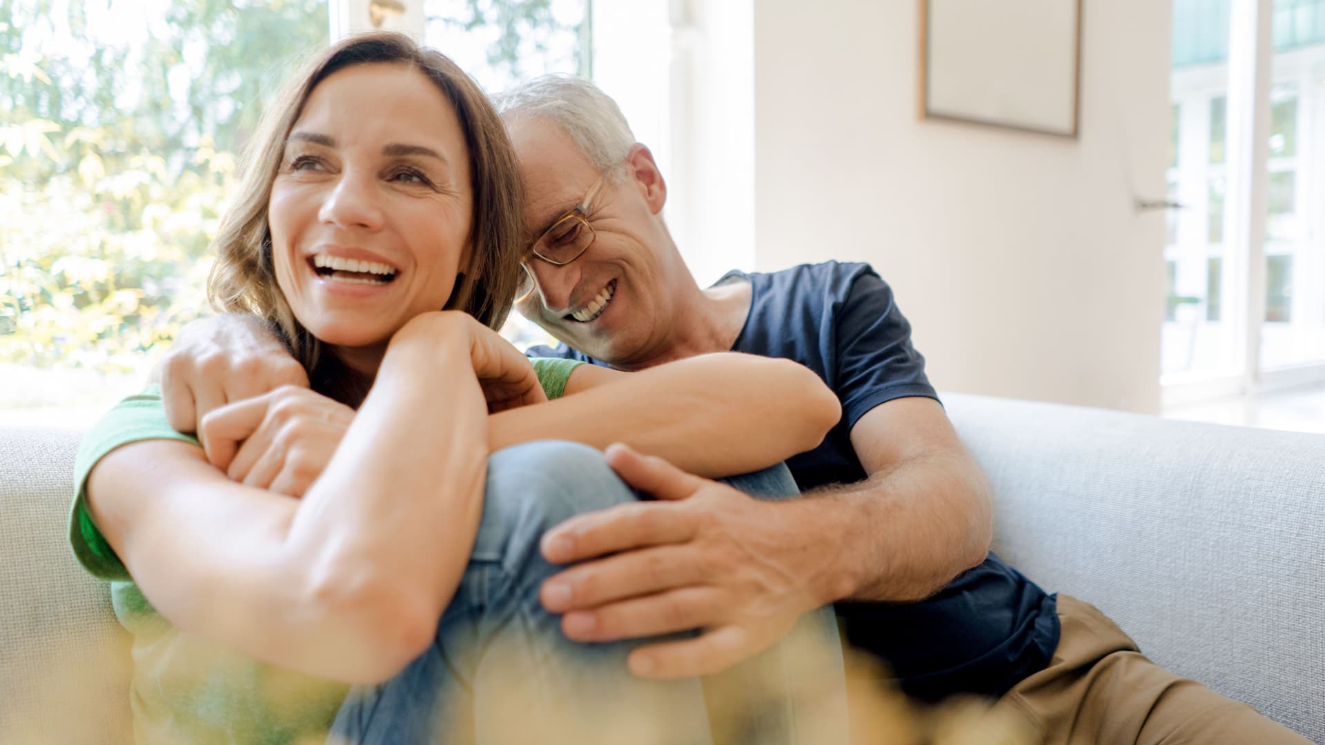 Lachendes reifes Paar sitzt auf der Couch zu Hause und umarmen sich.