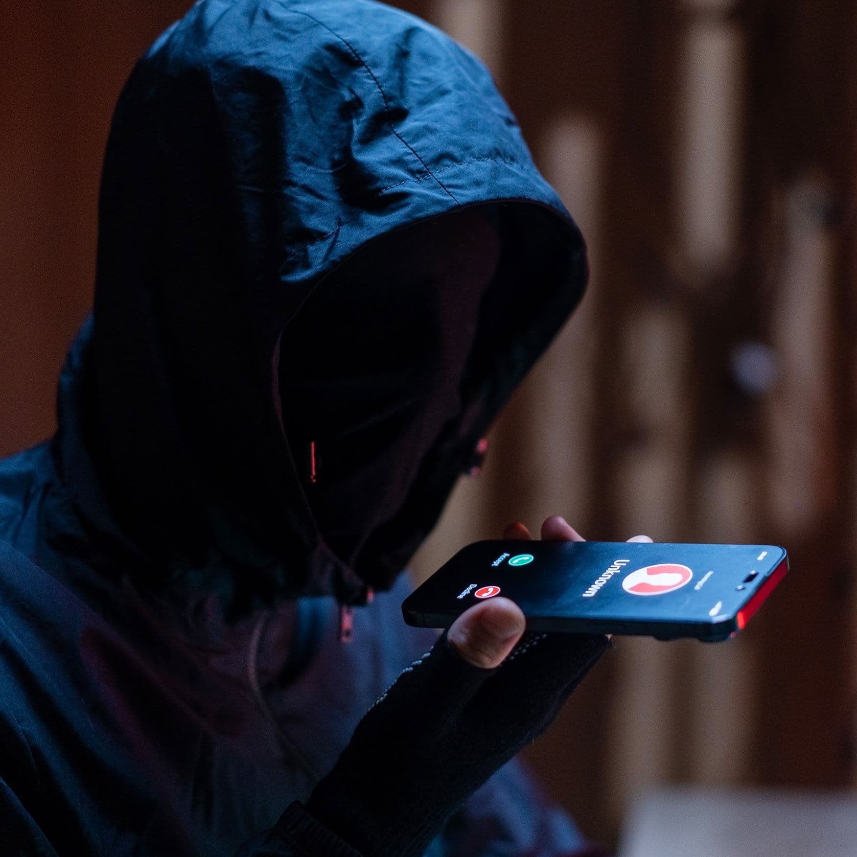 Person mit Kapuze und verdecktem Gesicht hält in dunkler Umgebung ein Smartphone mit eingehendem Anruf vor den Mund.