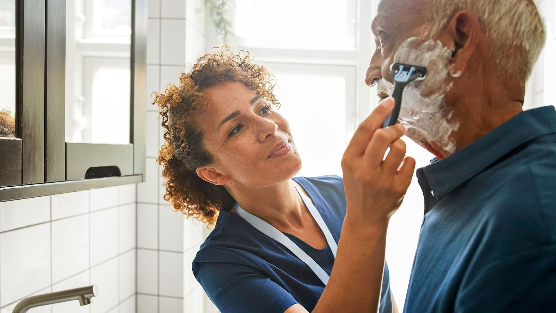 Eine Pflegerin hilft einem älteren Mann im Badezimmer beim Rasieren. Sie hält den Rasierer, während er mit Rasierschaum im Gesicht in einen Spiegel blickt. Die Szene wirkt fürsorglich und ruhig.