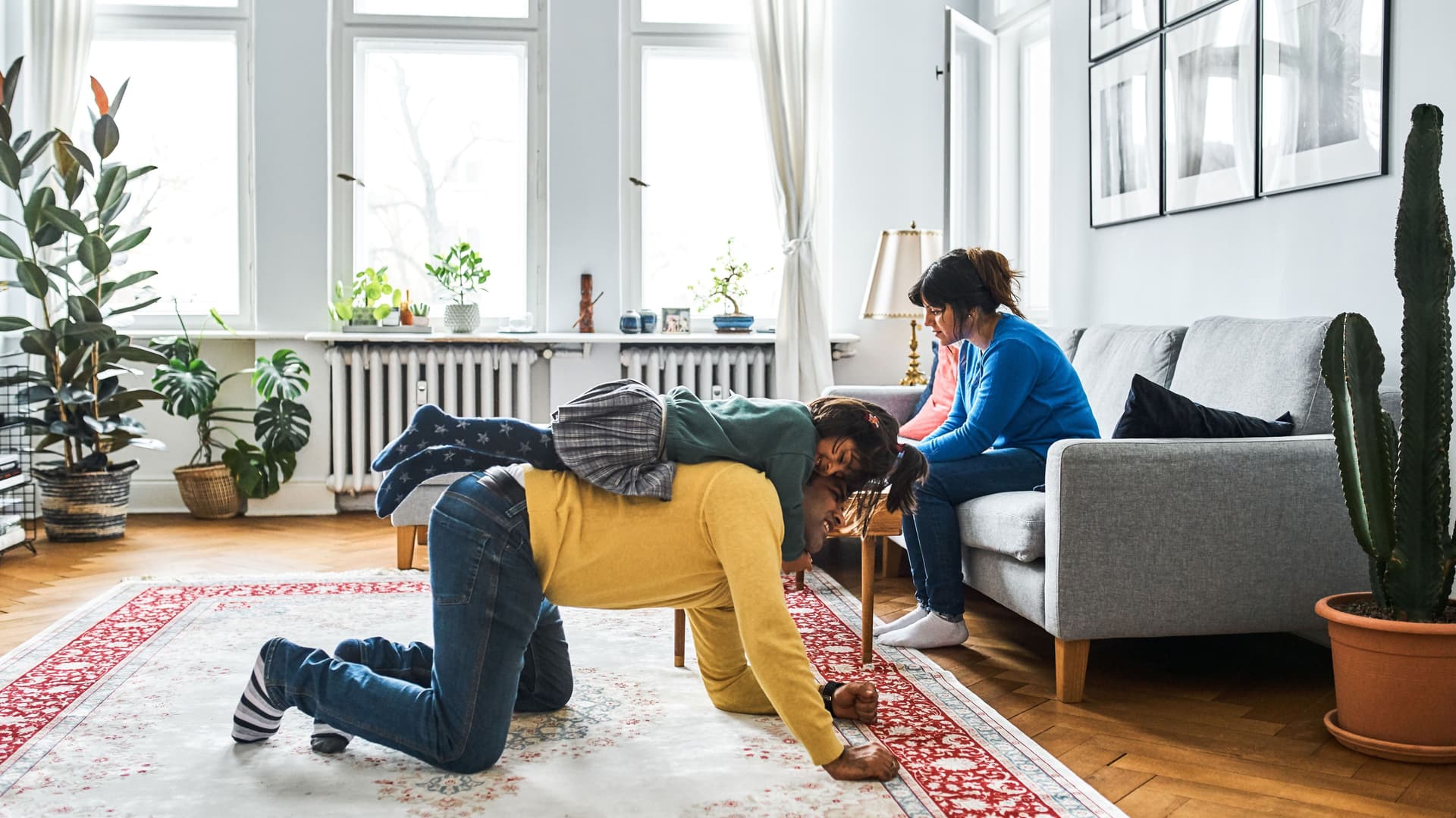 Ein Mann, der auf einem großen Teppich kniet, trägt ein kleines Mädchen auf dem Rücken. Eine Frau sitzt in dem großen hellen Altbauzimmer auf einem Sofa daneben.