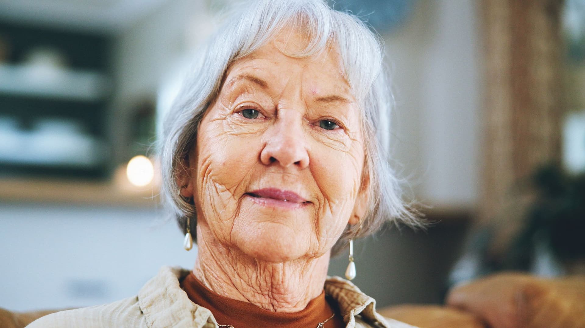 Eine Seniorin mit grauem, mittellangem Haar und freundlichem Gesichtsausdruck blickt direkt in die Kamera. Sie trägt Ohrringe und eine Kette. Der Hintergrund des Bildes ist unscharf, wodurch der Fokus ganz auf ihrem Gesicht liegt.