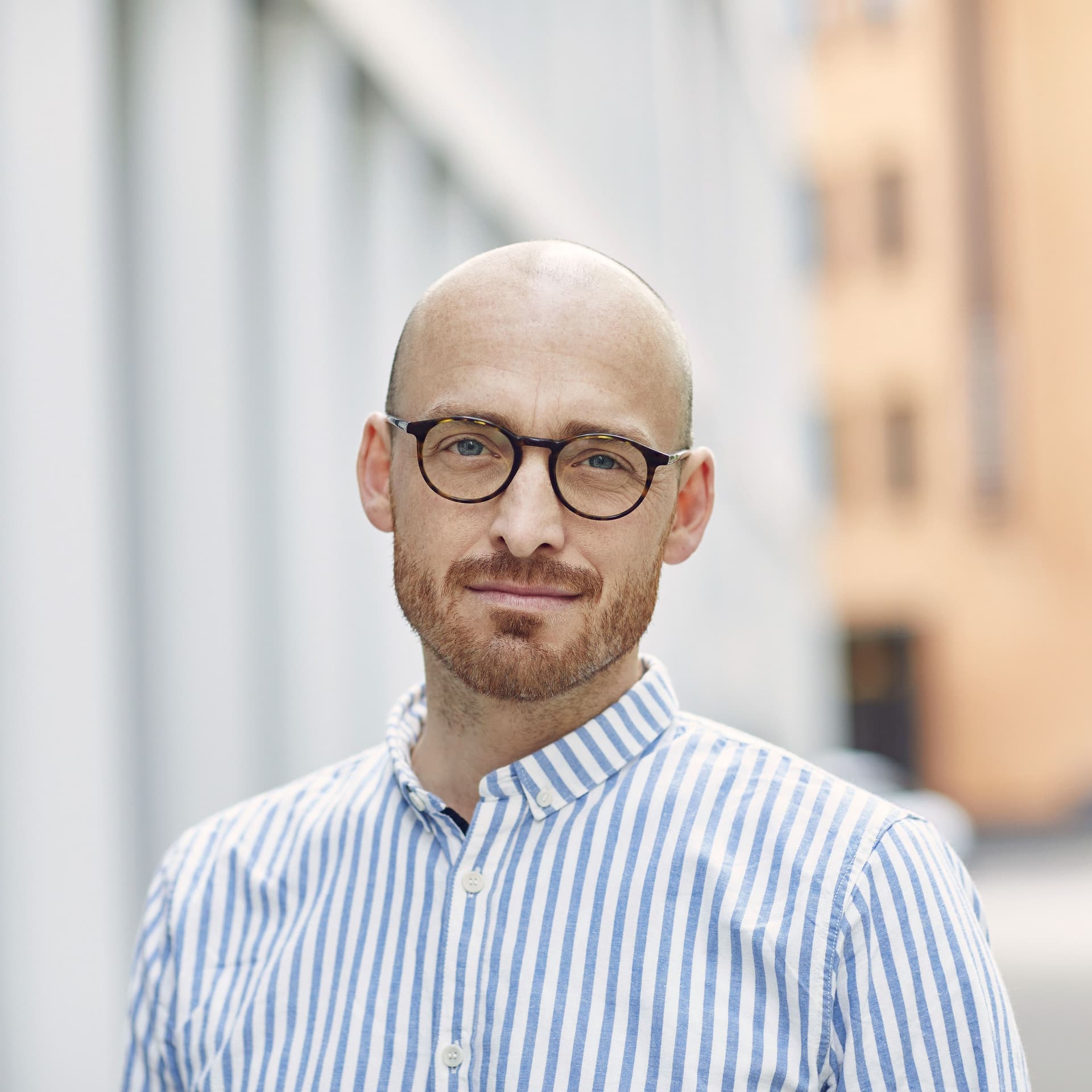 Porträt eines jungen Mannes mit gestreiftem Hemd. Er steht im Außenbereich vor Gebäuden.