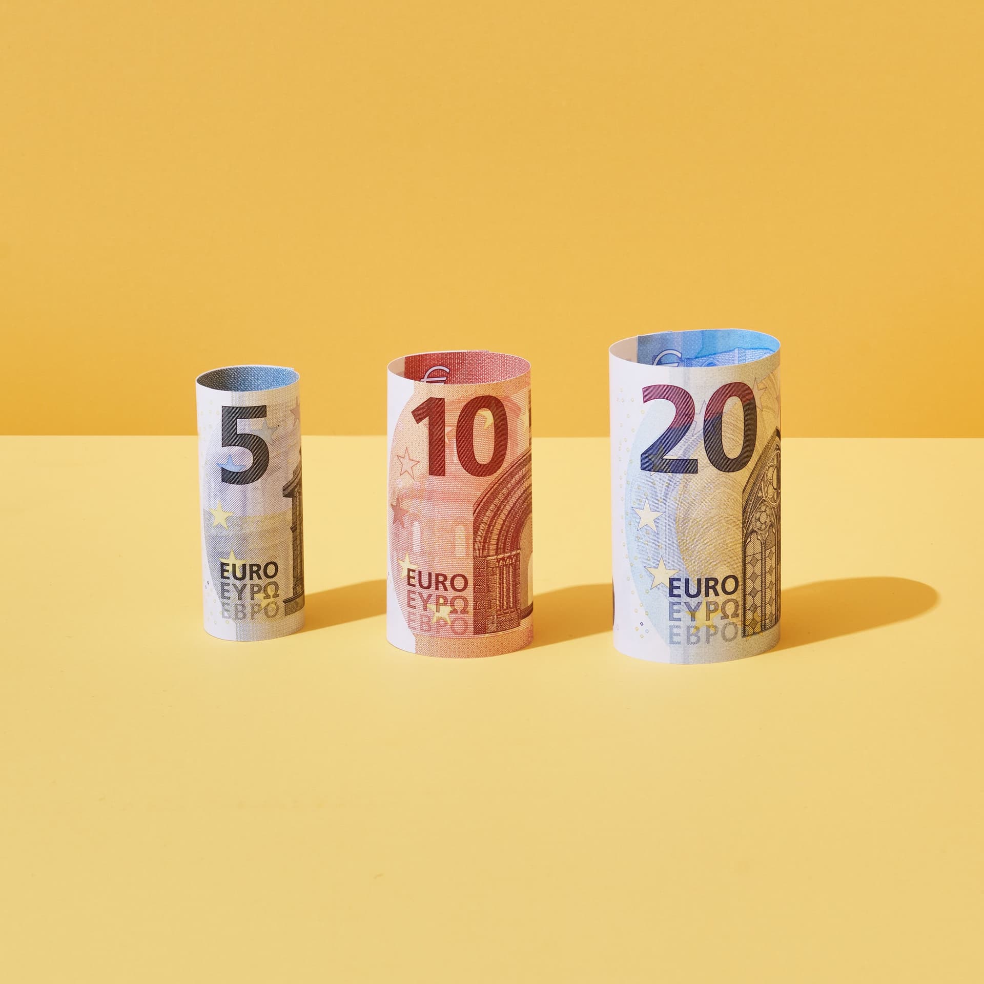 Jeweils ein gerollter 5-Euro, 10-Euro und 20-Euro-Schein stehen nebeneinander vor gelbem Hintergrund